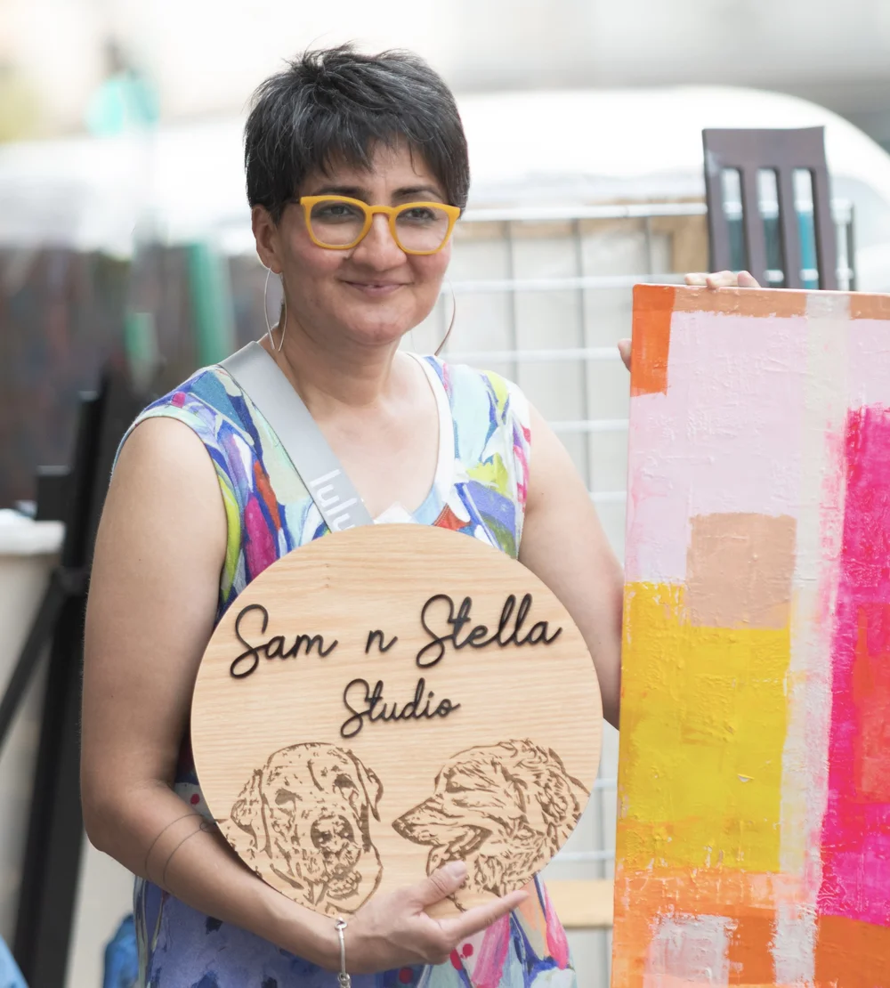 Rose holding her Sam n Stella Studio sign beside one of her abstract paintings